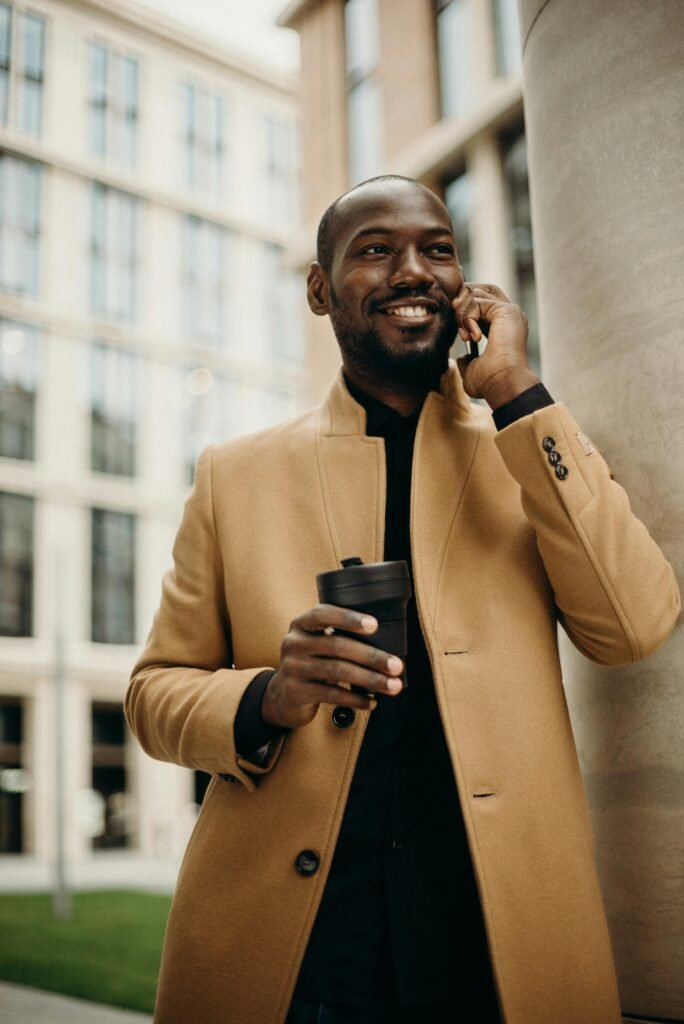 Smiling businessman in a trench coat holding a coffee cup while talking on the phone outdoors.